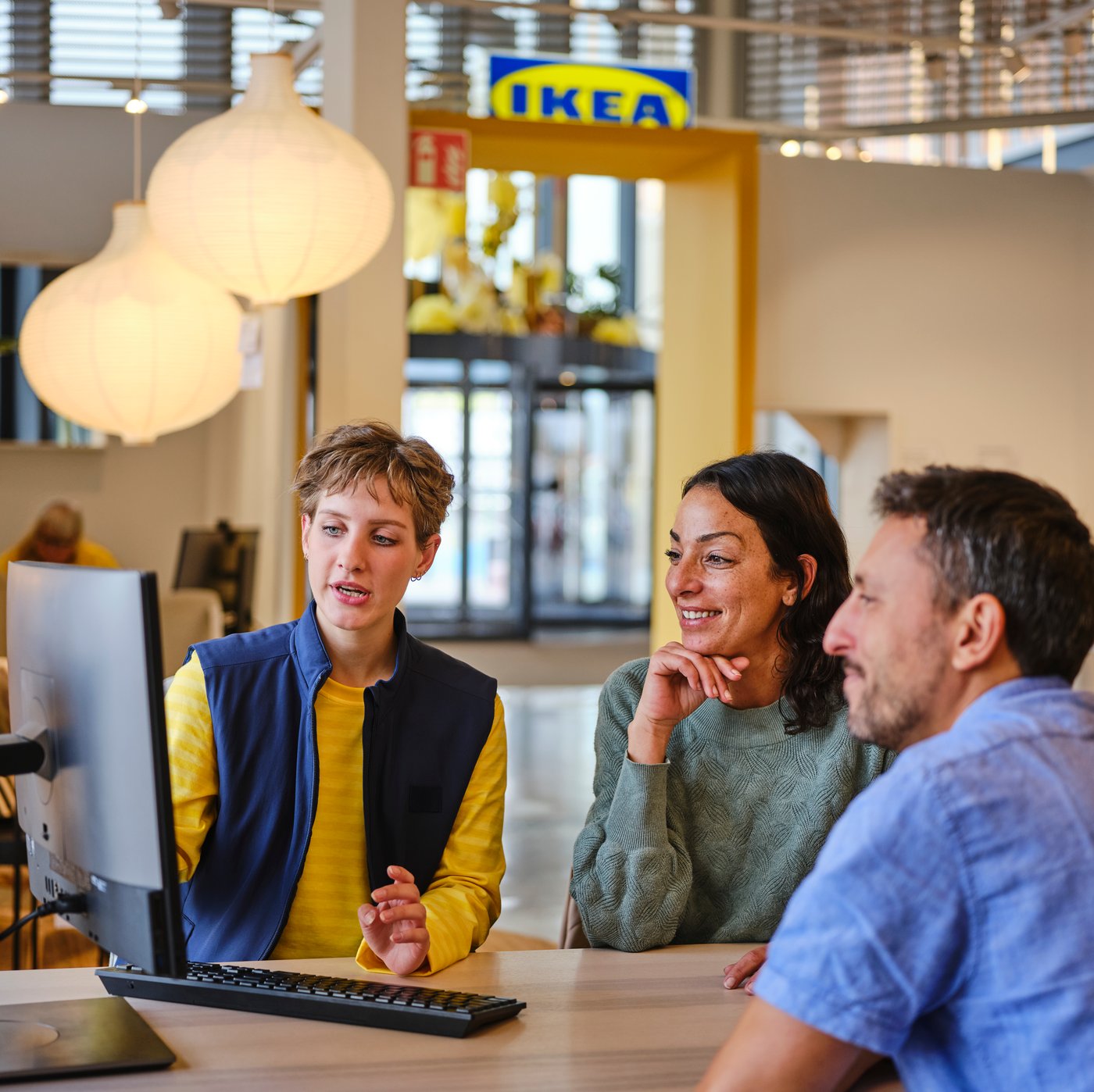 An IKEA co-worker wearing a uniform is talking with two customers in front of a computer.