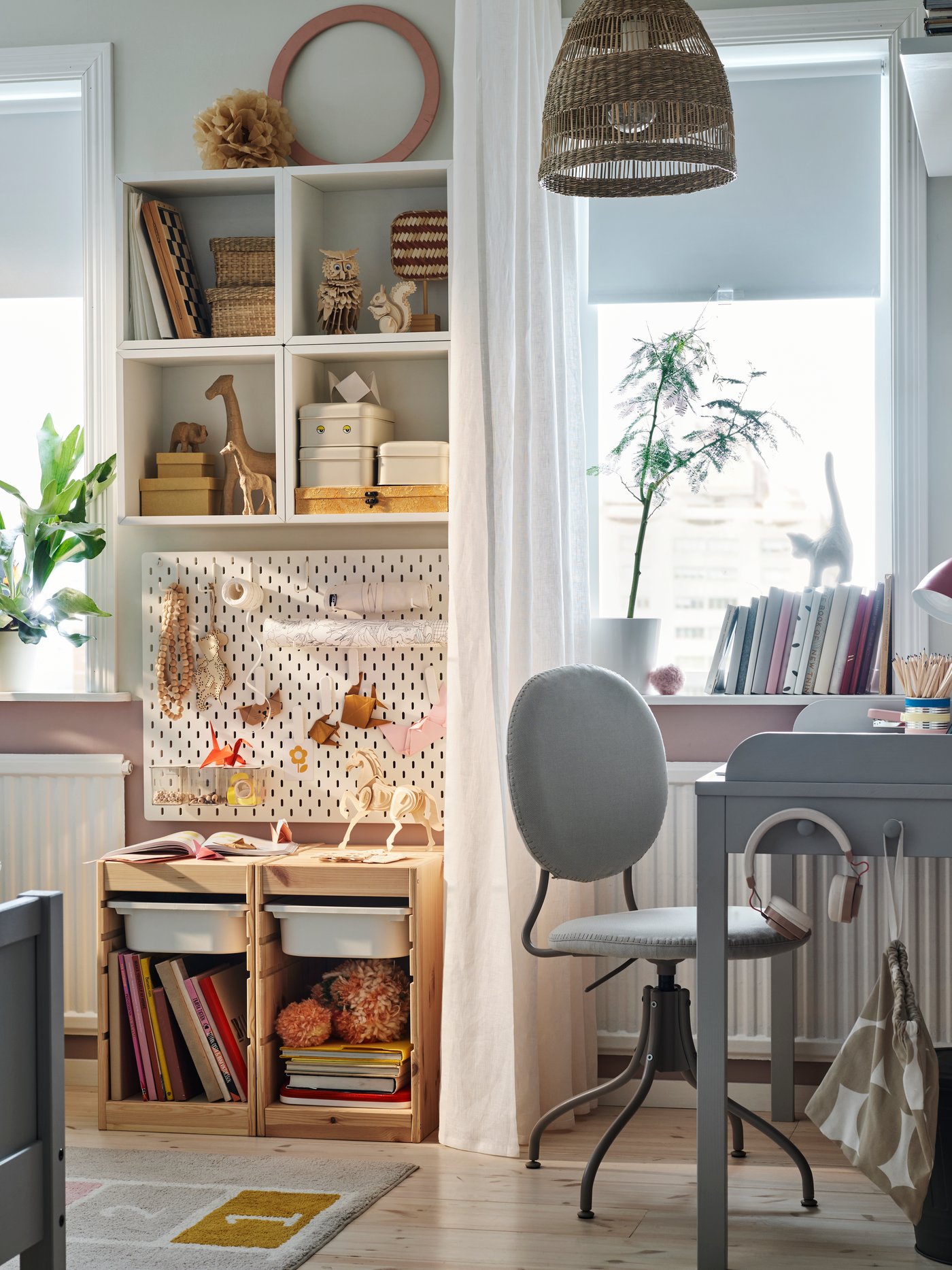 Knusse kinderslaapkamer met een grijs SMYGA bureau naast een raam afgewerkt met een wit gordijn. Naast het gordijn hangt een SKÅDIS ophangbord aan de muur.