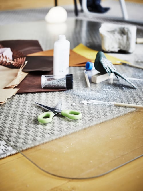 Drawing, painting and art supplies strewn across a plastic, transparent KOLON floor protector over a rug on a wooden floor.