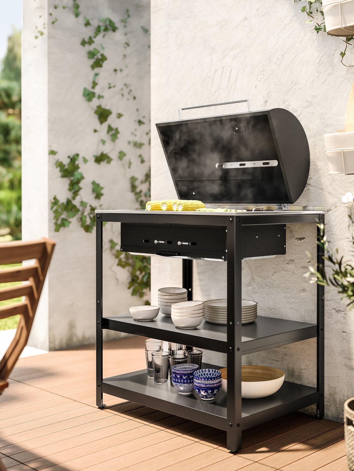 Ambiente al aire libre con una barbacoa de carbón con cuencos y vasos en los estantes inferiores. La barbacoa está en el suelo.