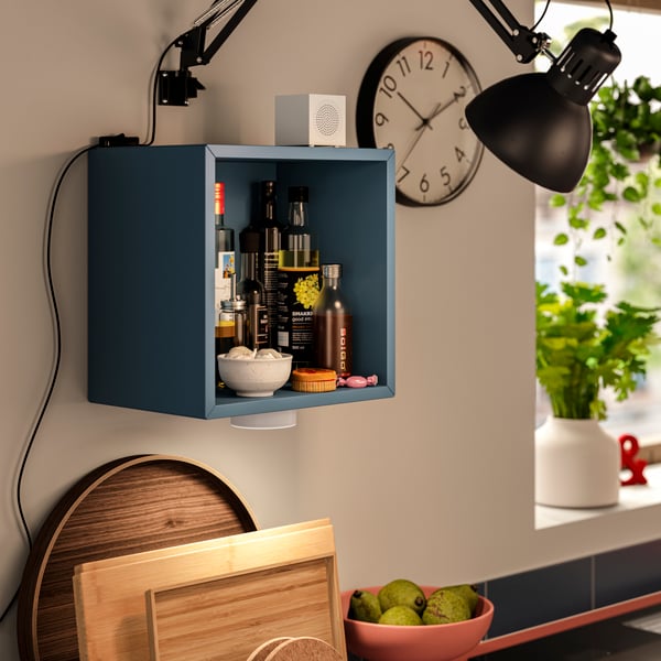 A dark grey-blue EKET wall-mounted shelving unit is placed on a kitchen wall and has bottles and jars stored inside it.