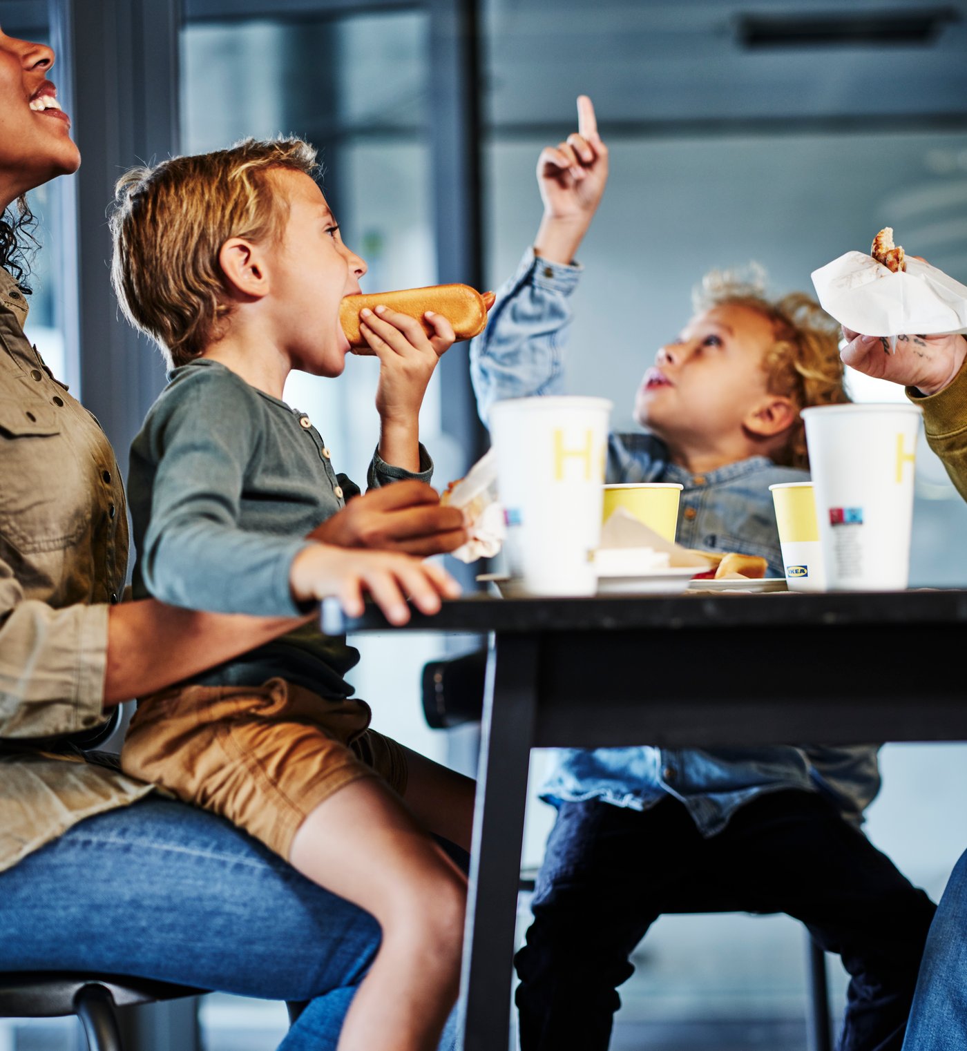테이블에 앉아 핫도그와 음료를 먹으며 즐겁게 식사하는 아이들과 보호자의 이미지