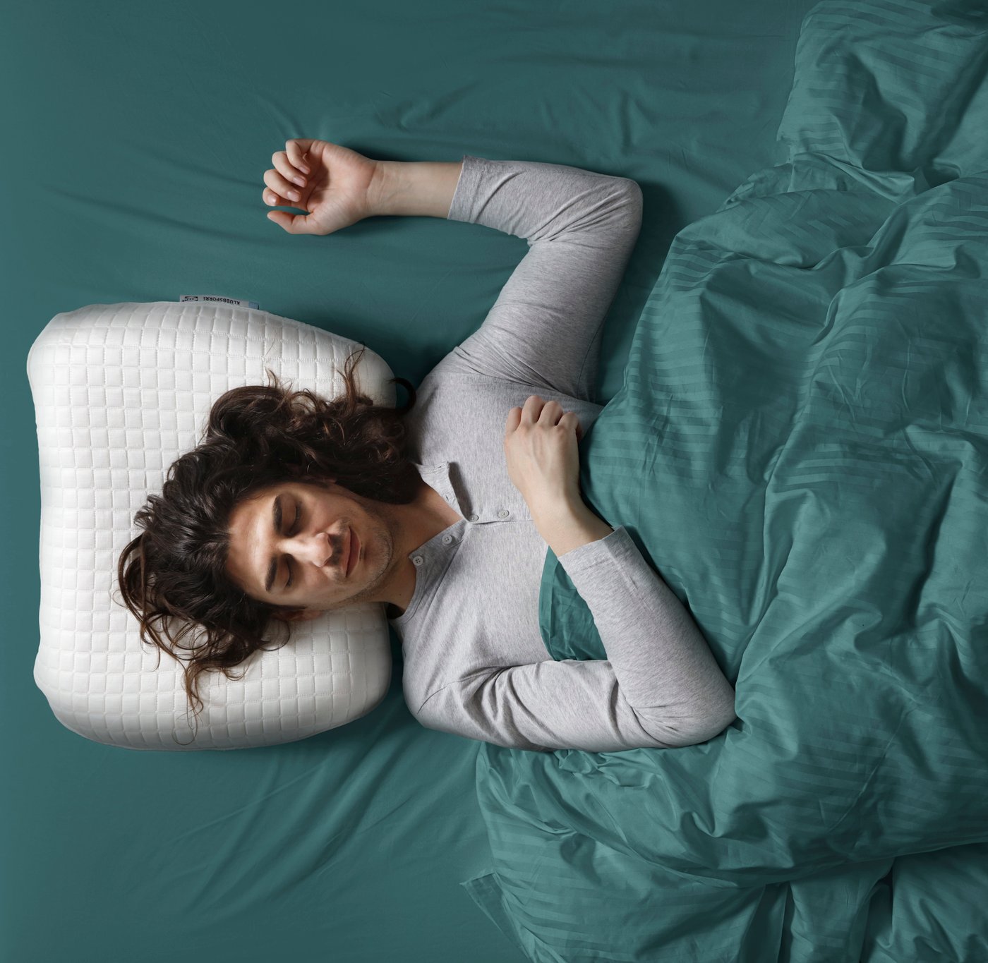 Vogelperspektive einer Person, die auf dem Rücken schläft, mit erhobenem Arm und dem Kopf auf einem ergonomischen KLUBBSPORRE-Kissen.