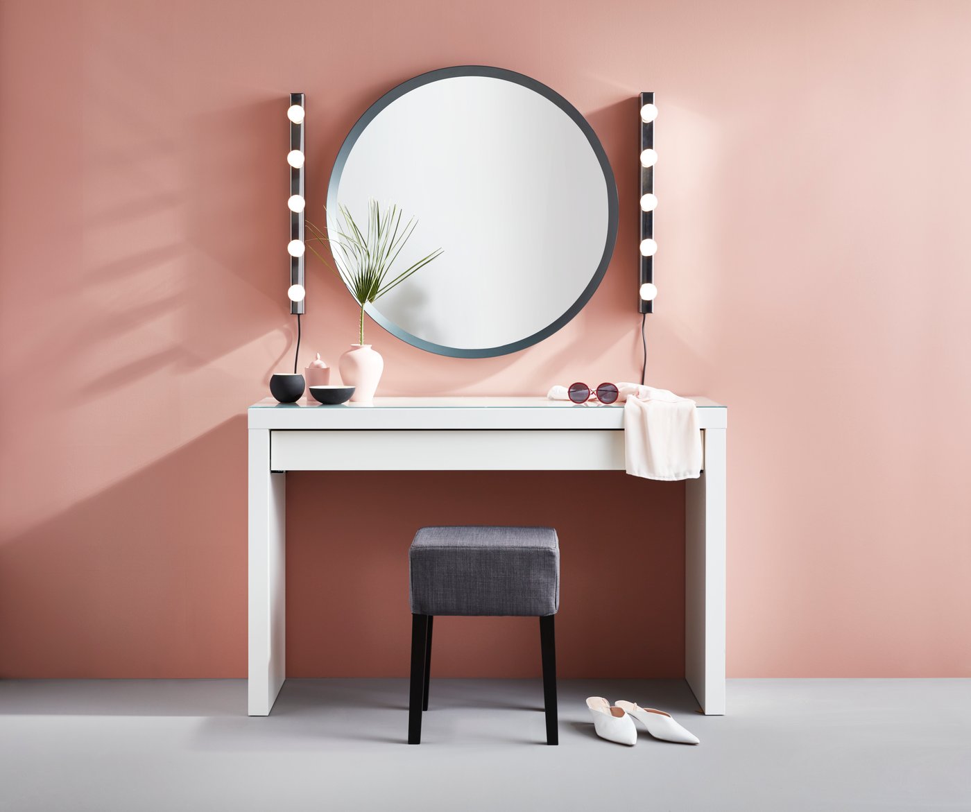En sminkstaion med spegel och belysning vid rosa vägg. 
