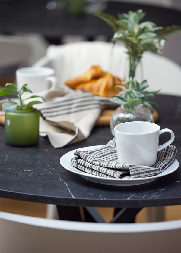 Närbild på runt bord med svart marmorskiva dukat med vit kopp på tallrik, tygservett, liten grön växt i kruka och croissanter på skärbräda i trä.