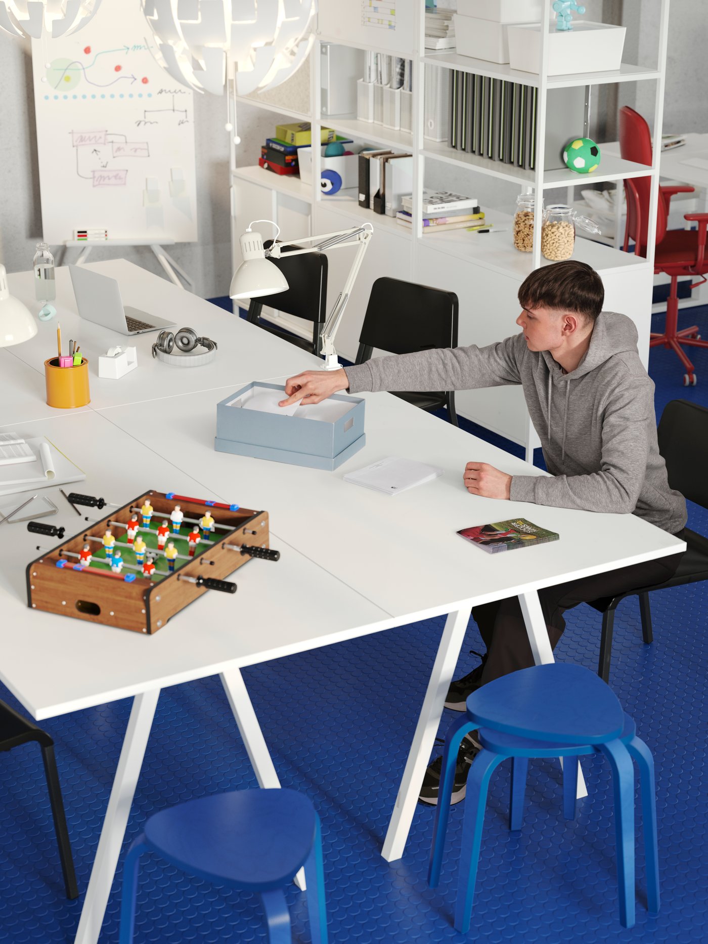 A meeting area of four white TROTTEN desks, black chairs and blue stools. A boy is folding flyers wearing a grey sweater.