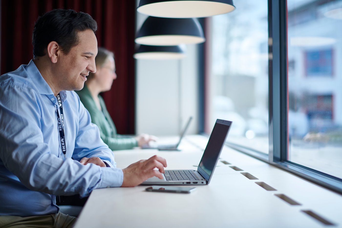 Une personne est assise à un bureau devant un ordinateur portable ouvert, face à une grande fenêtre.