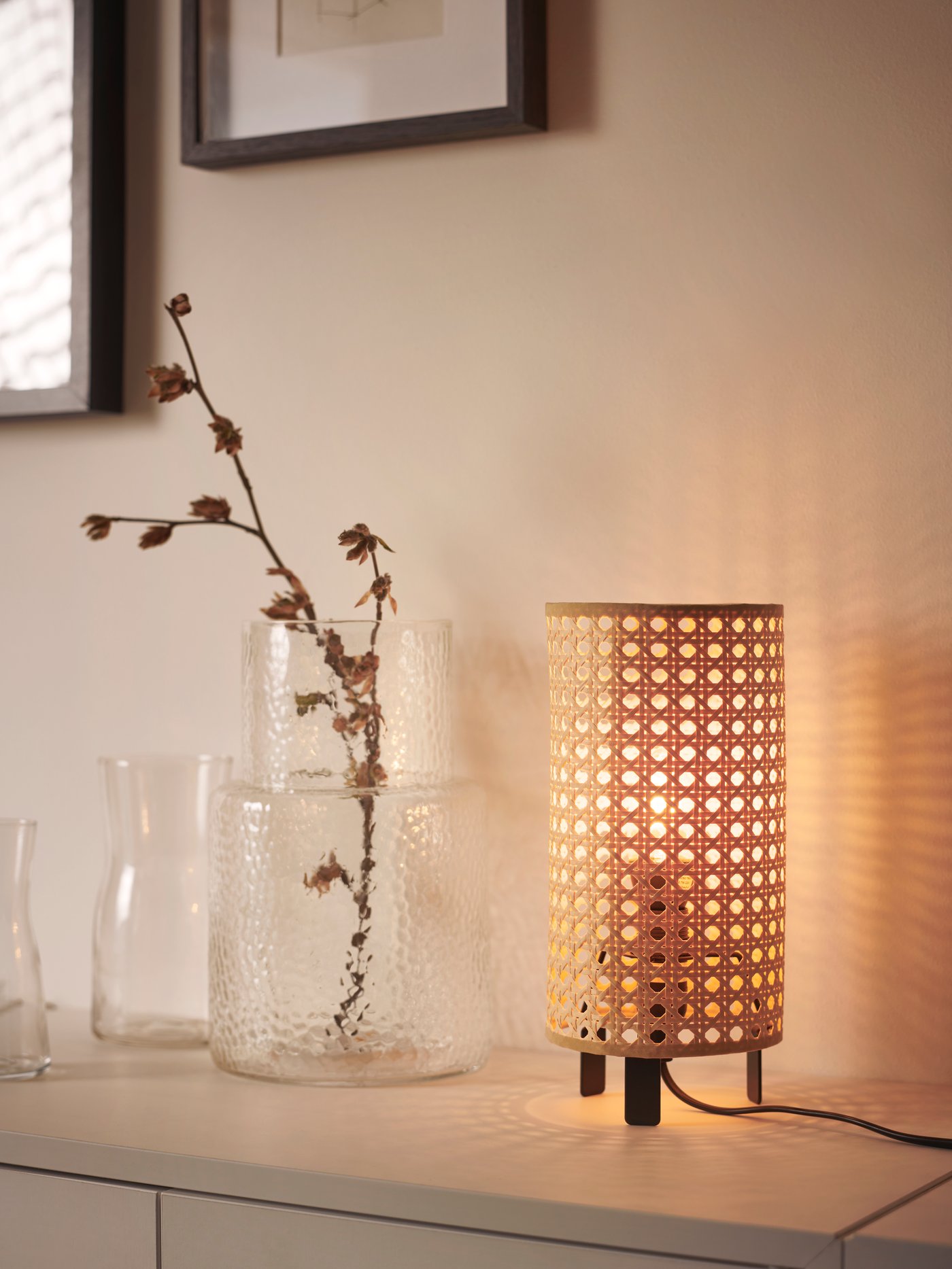 Lampe allumée sur une commode blanche à côté d'un vase transparent contenant une plante