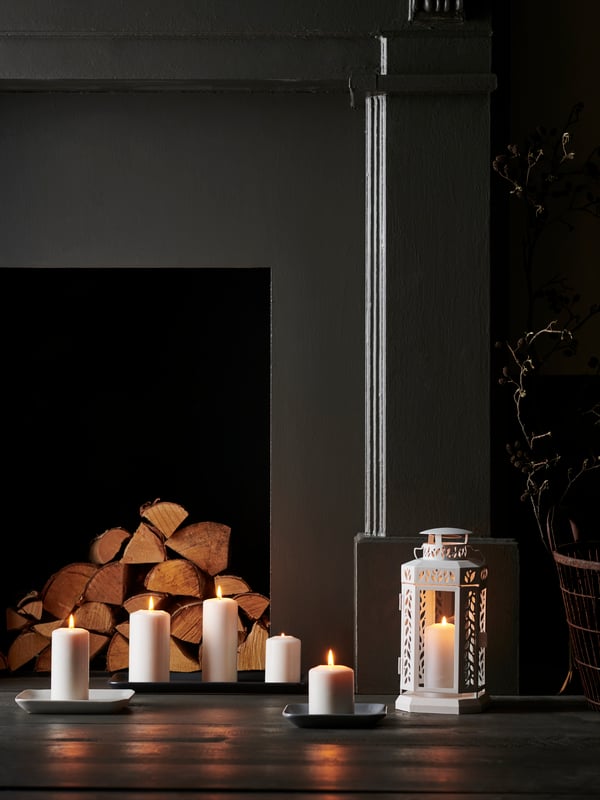 Un camino nero con legna impilata all'interno. Di fronte, ceri bianchi accesi FENOMEN davanti a una lanterna bianca.