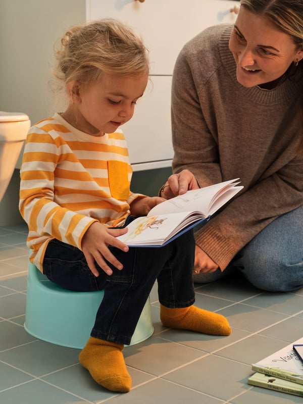 A small child sitting on LILLA children’s potty, reading a book and with the mother sitting by the side to help.