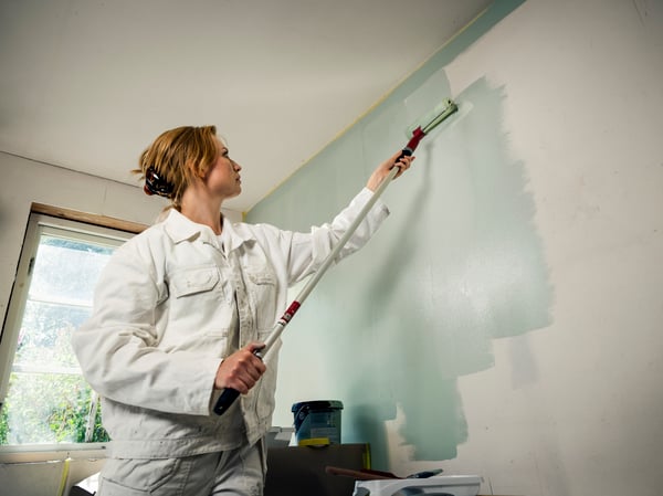 A person in white overalls uses a roller to paint a wall light blue in a brightly lit room.