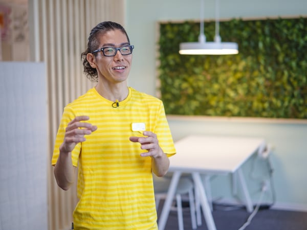 眼鏡をかけた黄色のストライプシャツの人物が、緑のある室内で話している