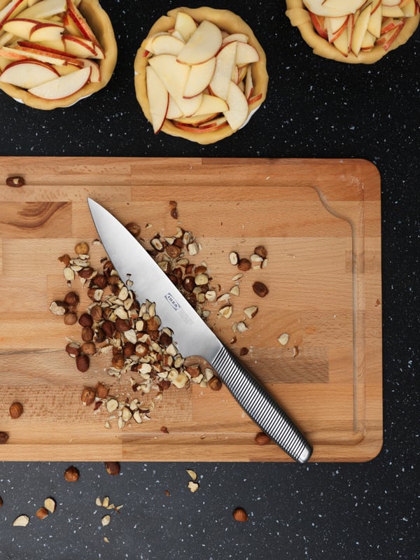 An IKEA 365+ cook’s knife of stainless steel is placed on a wooden chopping board with a fistful of nuts, partly chopped.