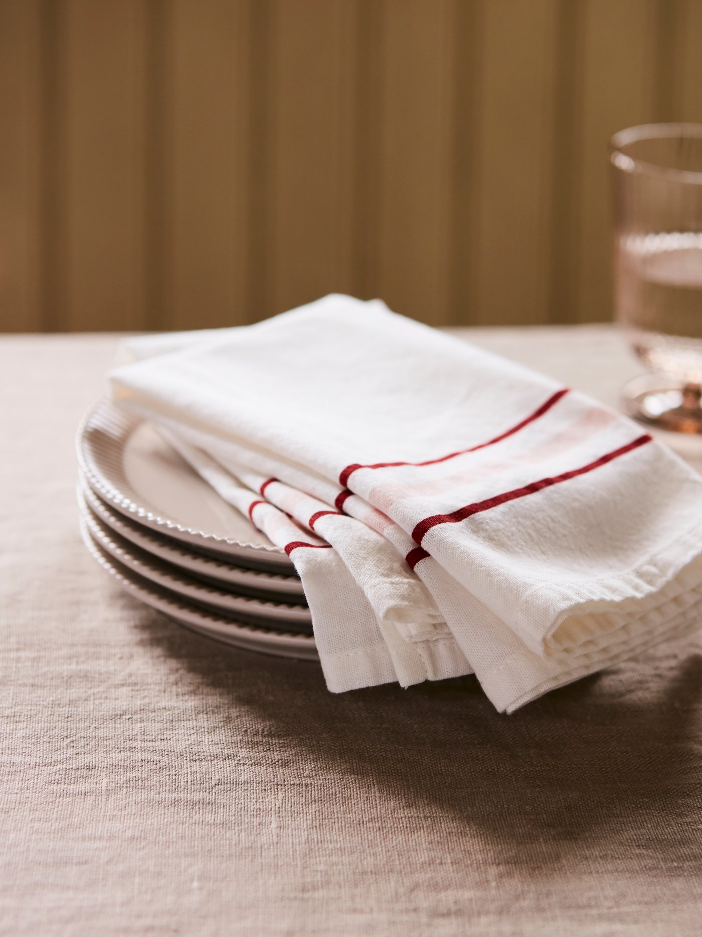 Deux serviettes de table en tissu ANLEDNING blanches ornées de rayures rouge et rose sur le bord reposent sur une pile d’assiettes sur une table.