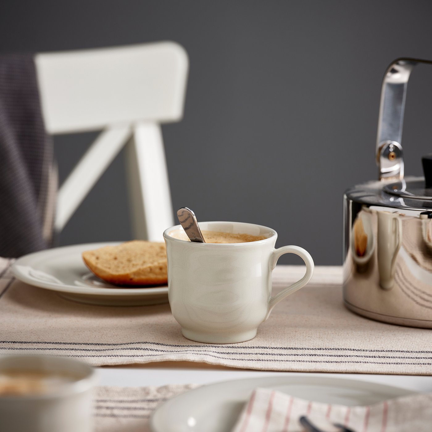 A SANDSKÄDDA mug in light grey-beige with a spoon inside it is standing next to a kettle and a plate with some bread on it.