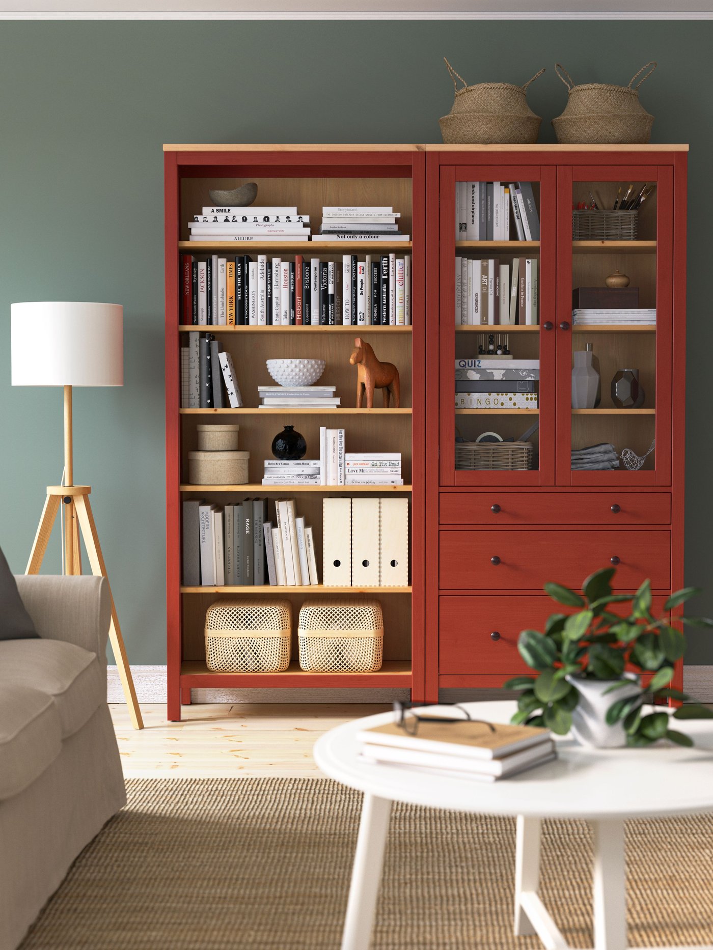 Červeno-dřevěná knihovna v obýváku, s otevřenými policemi a prosklenými dvířkami a zásuvkami. Knihovna je plná knih, dekorací a organizerů, a vedle ní stojí bílá lampa s dřevěnými nohami.