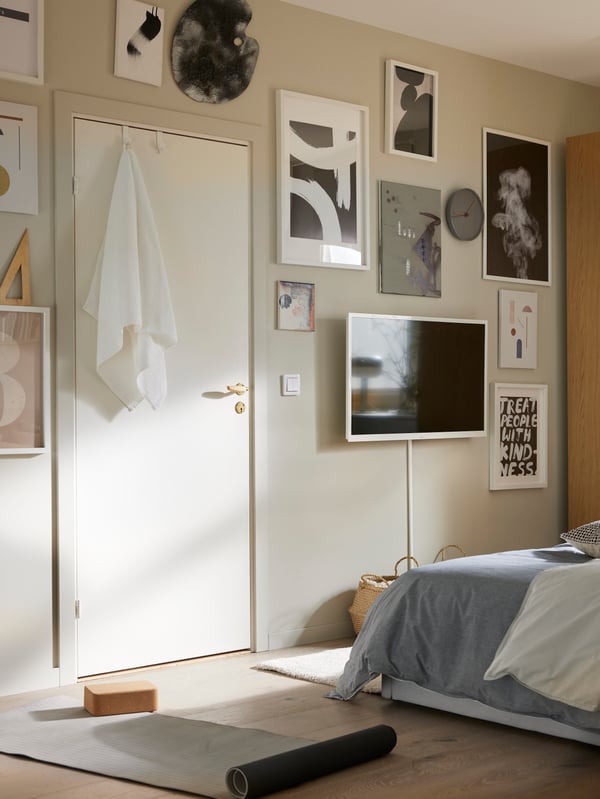 Una collezione di poster BILD con cornici bianche e un TV su una parete beige, di fronte a un letto con un piumino HEDFRYLE.