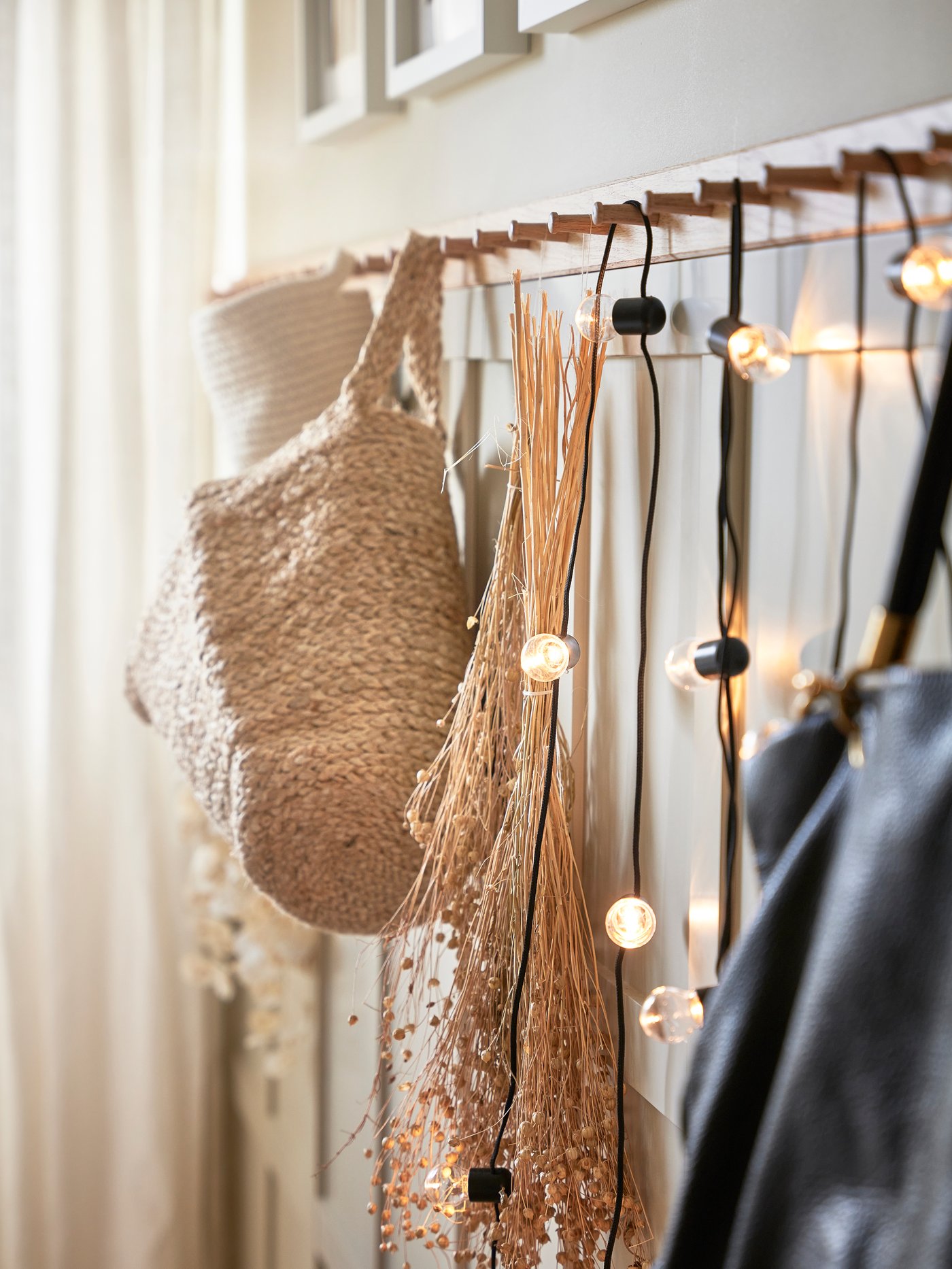 En rad med krokar där väskor och nätkassar i naturmaterial hänger, dekorerat med små ljusslingor framför ljusa gardiner.
