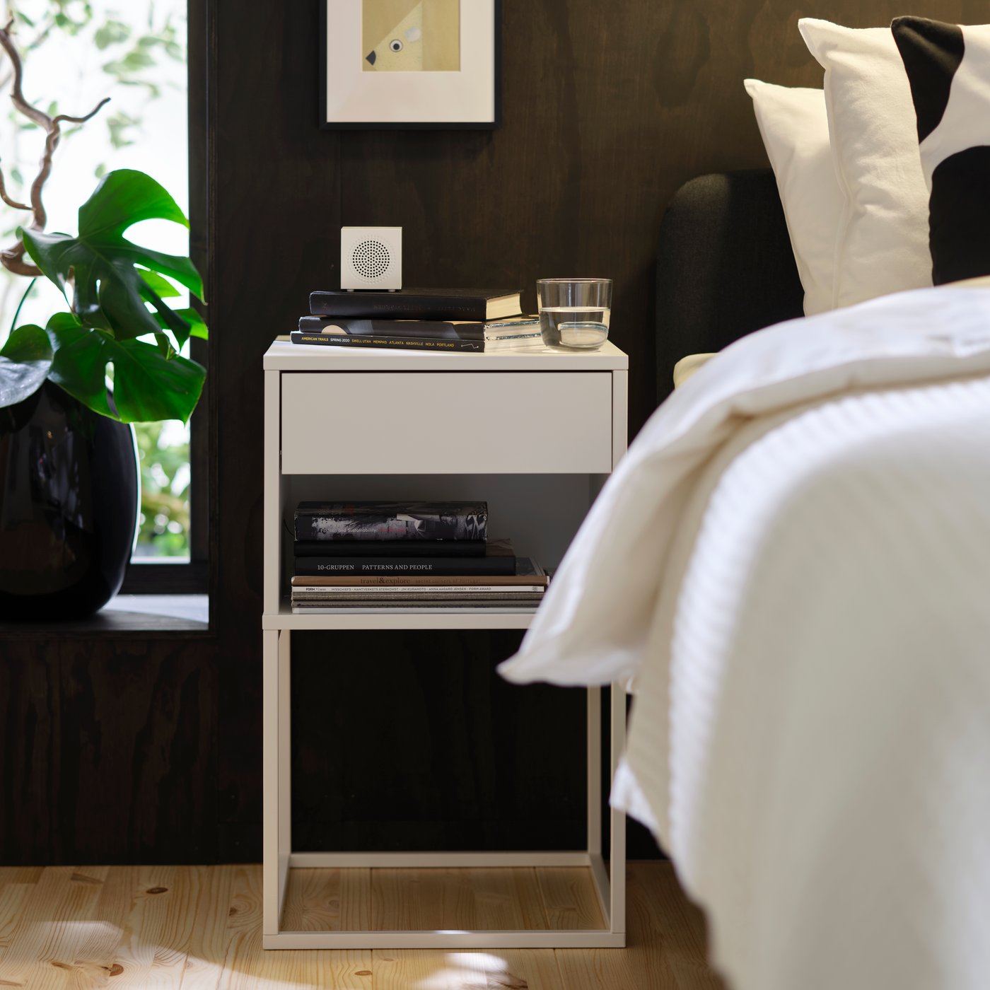 HATTÅSEN bedside table in white with drawer and shelves holding books and glass placed beside a bed in a dark-toned bedroom.