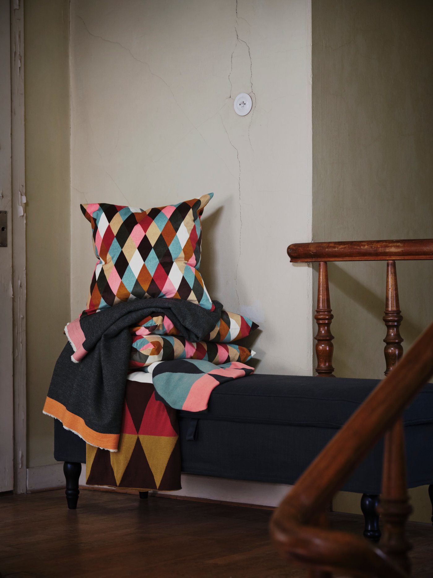 Diamante-eredua eta kolore biziak dituzten DEKORERA tapaki eta kuxinak banku baten gainean pilatuta, eskailera baten ondoan.