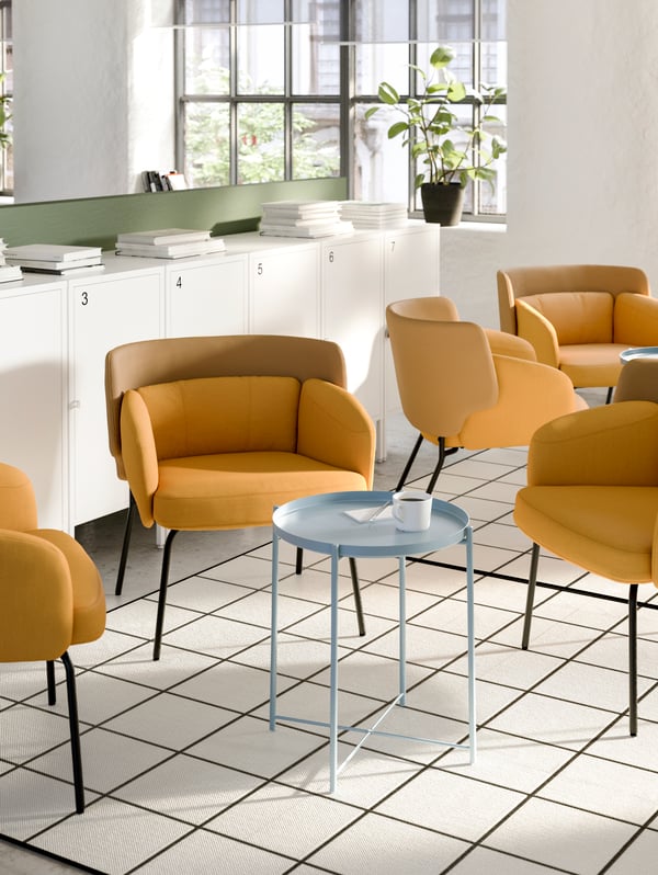 A lounge area with two groups of yellow armchairs, small round metal tables in the middle and a row of windows behind.