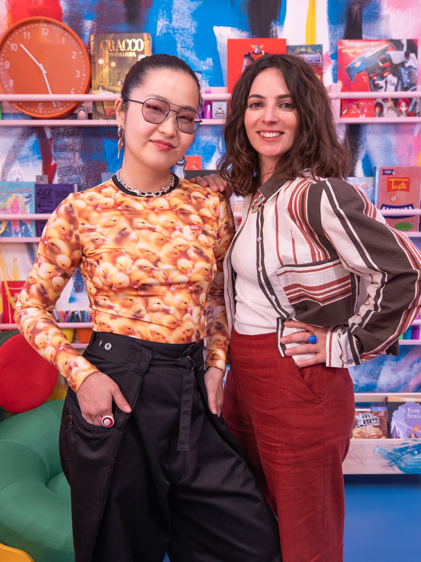 Lydia Chan e Alessandra Lauria, coppia creativa per la Milano Design Week 2026, in posa fianco a fianco in un ambiente colorato, con scaffali pieni di libri e oggetti decorativi sullo sfondo.