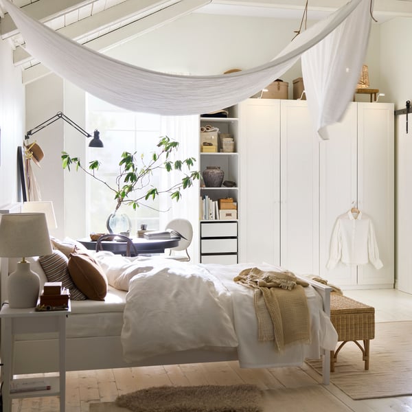 A white HEMNES bed covered in white DYTÅG bed linen and brown cushions with a DYTÅG curtain draped from the ceiling above it.