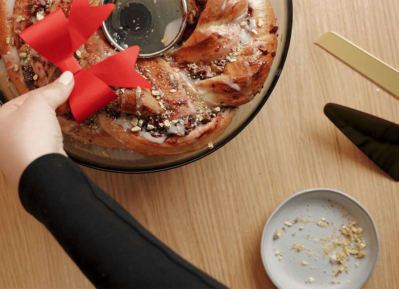 Eine Hand legt ein rotes Band auf einen festlichen, glasierten Gebäckkranz mit Nüssen. In der Nähe liegen ein goldener Tortenheber und ein kleiner Teller mit Krümeln auf einem Holztisch.