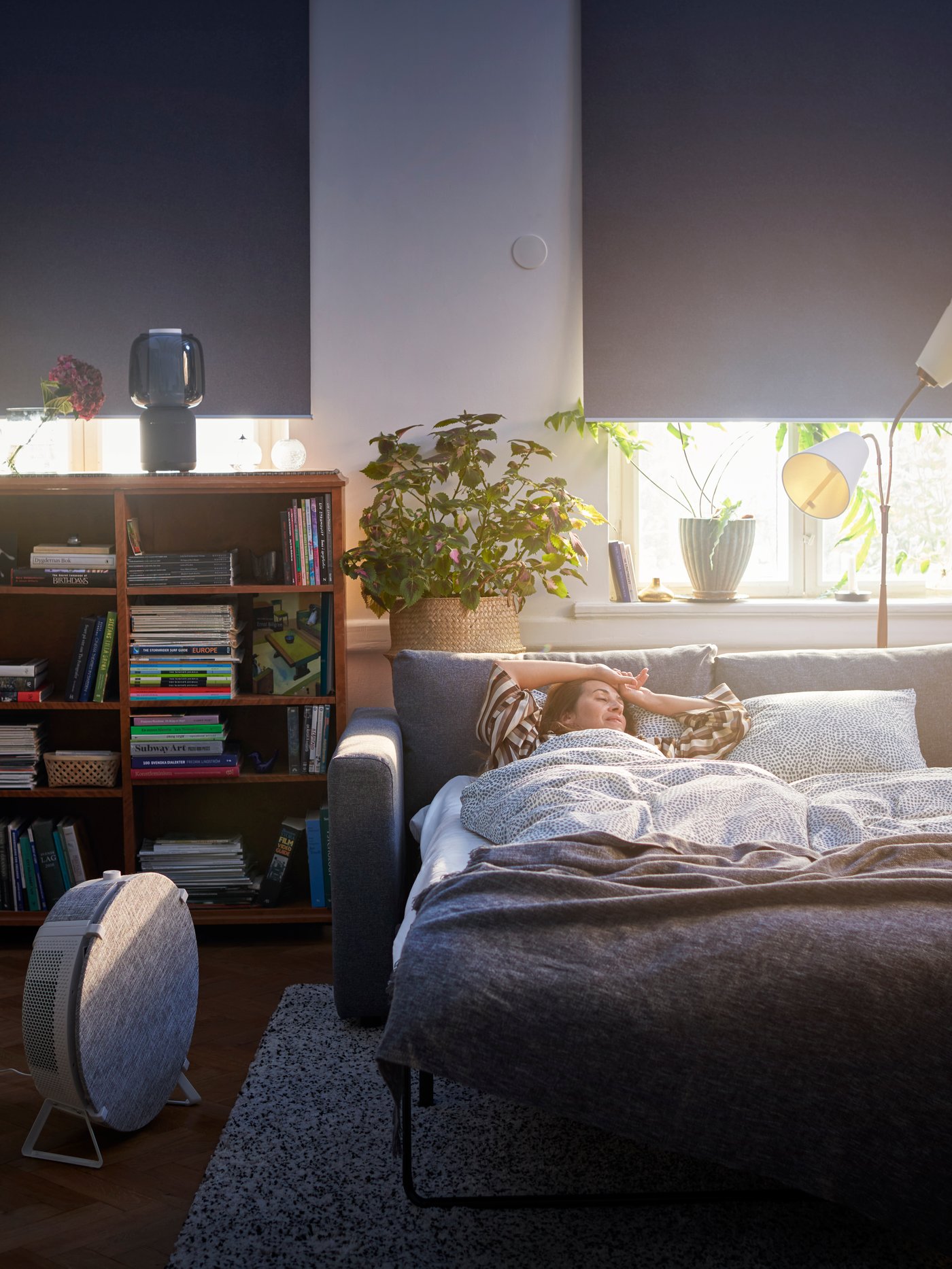 Un divano letto VIMLE sfondato con una donna che dorme sopra e una lampada con altoparlante SYMFONISK nera con Wi-Fi e paralume in vetro accanto.