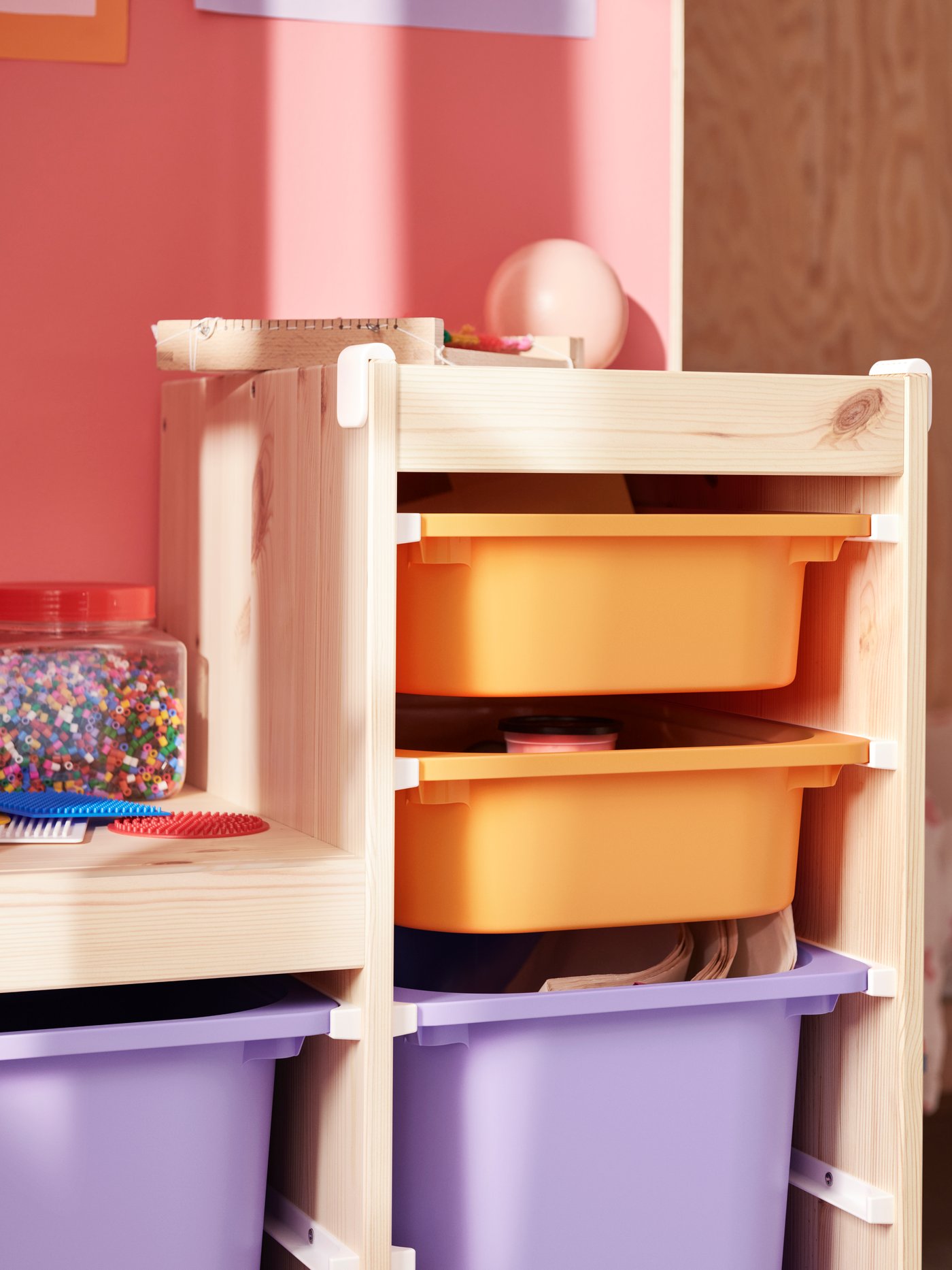A wooden shelf with colorful TROFAST plastic bins — two orange and one purple — in a room with pink walls.
