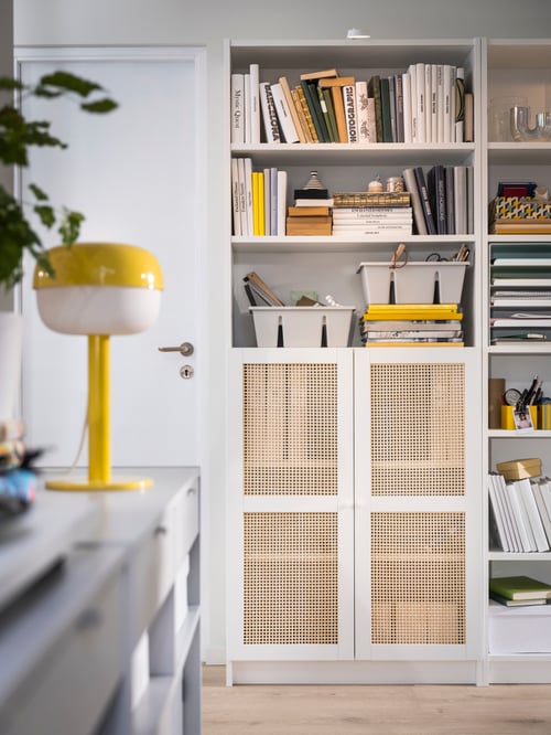 Two HÖGADAL doors placed on a bookcase filled with books and an LED cabinet lighting hanging on it, and a table lamp nearby.