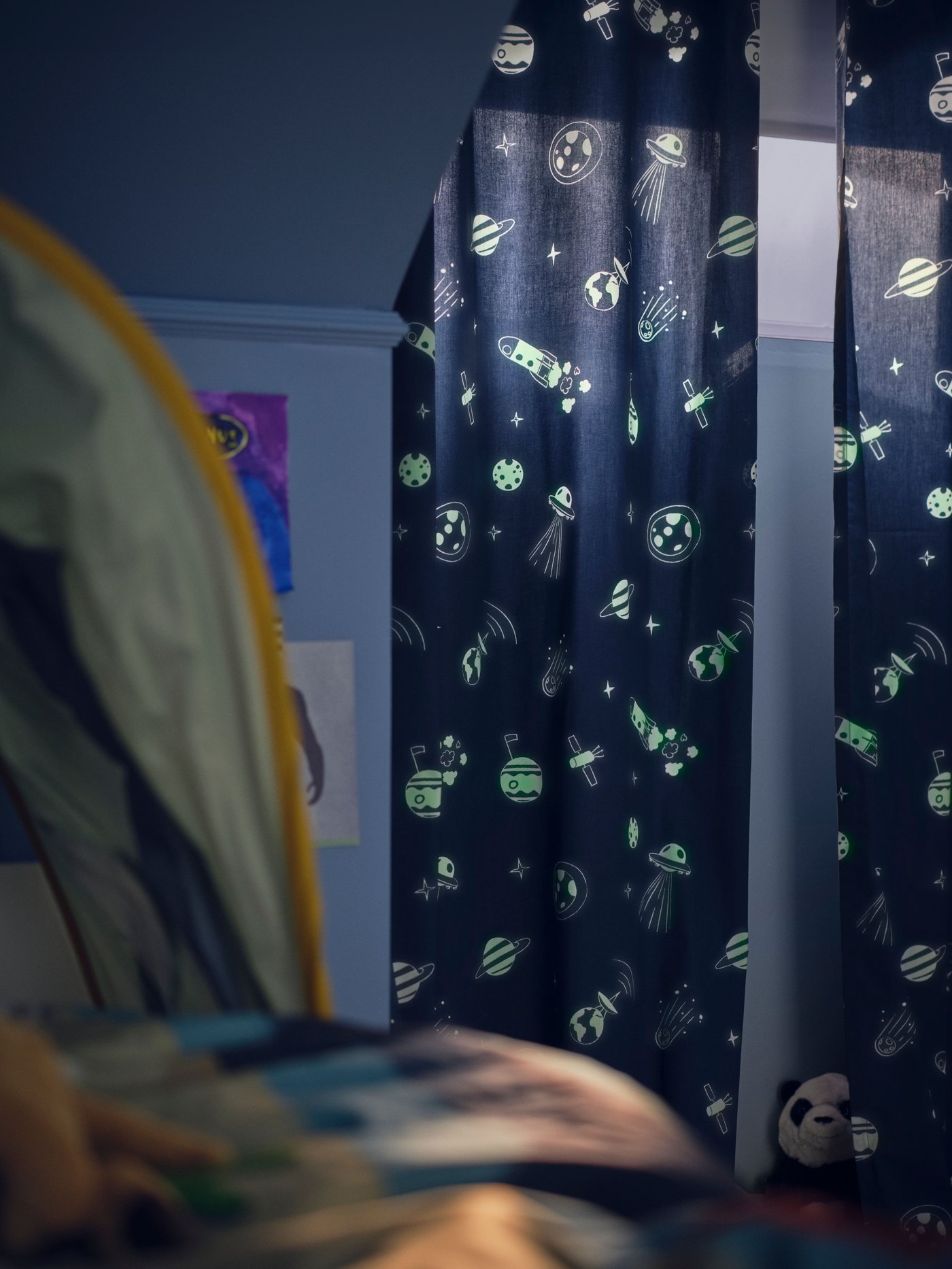 A dim blue children’s bedroom with an AFTONSPARV glow-in-the-dark curtain across the window, showing glowing space motifs.