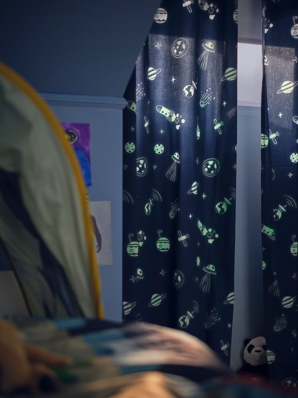 A dim blue children’s bedroom with an AFTONSPARV glow-in-the-dark curtain across the window, showing glowing space motifs.