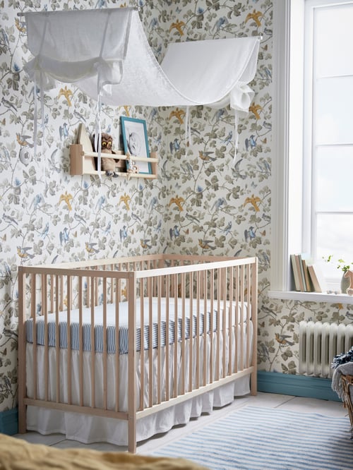 Un lit pour enfant SNIGLAR en hêtre est posé dans le coin d’une pièce tapissée de papier peint. Il est doté de linge de lit à rayures bleues et d’une jupe de lit blanche.