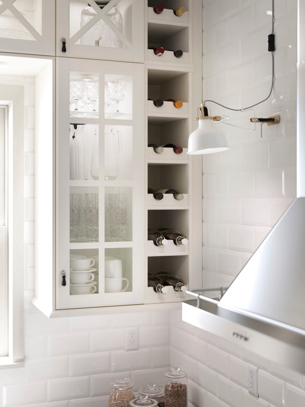 Cabinets with off-white glass doors on a white brick wall, and open cabinets with slots to store bottles on their side.