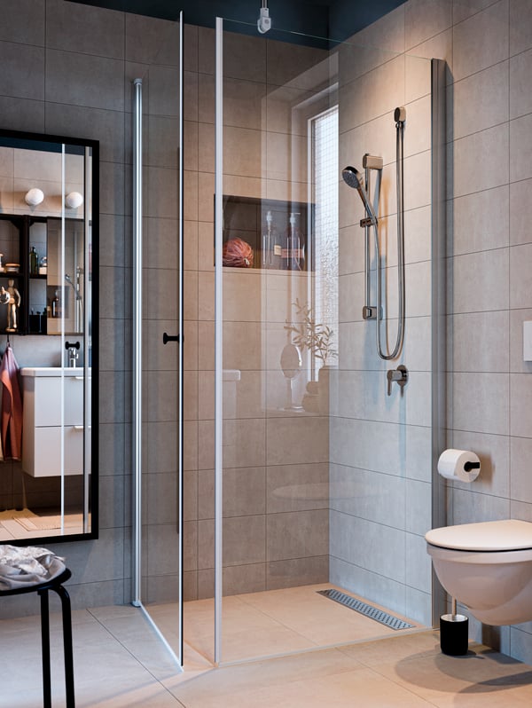 Modernes IKEA-Badezimmer mit begehbarer Glasdusche, stilvoller Regendusche und eleganten, hellgrauen Wandfliesen. Im Spiegel ist ein weiterer Blick auf das geschmackvoll eingerichtete Badezimmer mit IKEA-Badmöbeln und dezenten Dekoelementen zu sehen