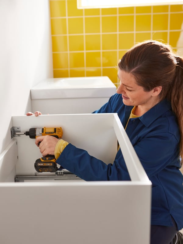 Une femme vêtue d’un uniforme IKEA installe un meuble pour lavabo dans une salle de bains jaune vif.
