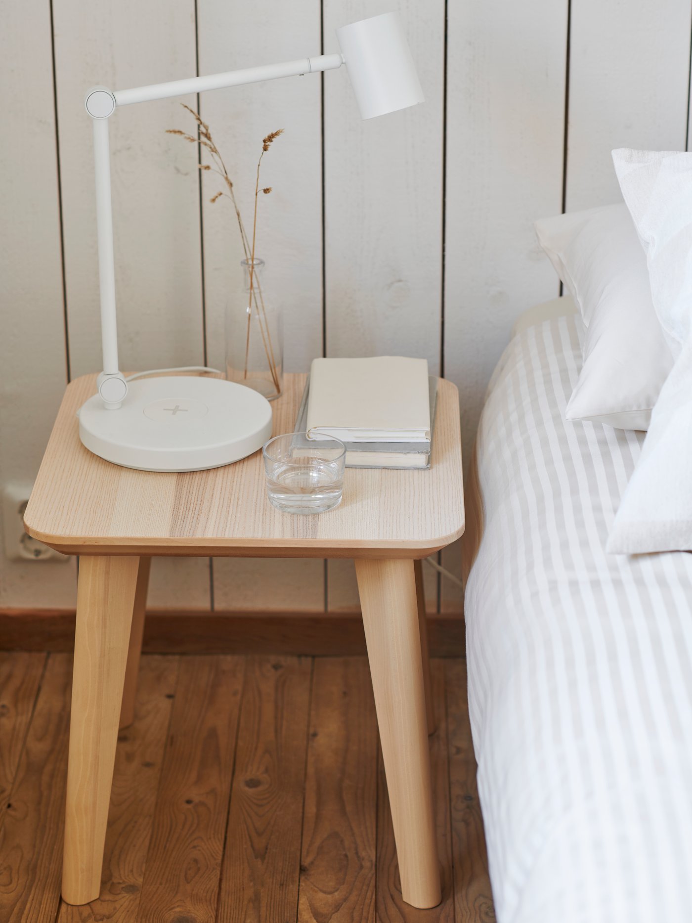 طاولة جانبية LISABO عليها مصباح مكتب NYMÅNE وكوب ماء وكتب، بجوار سرير في غرفة نوم على الطراز الريفي.