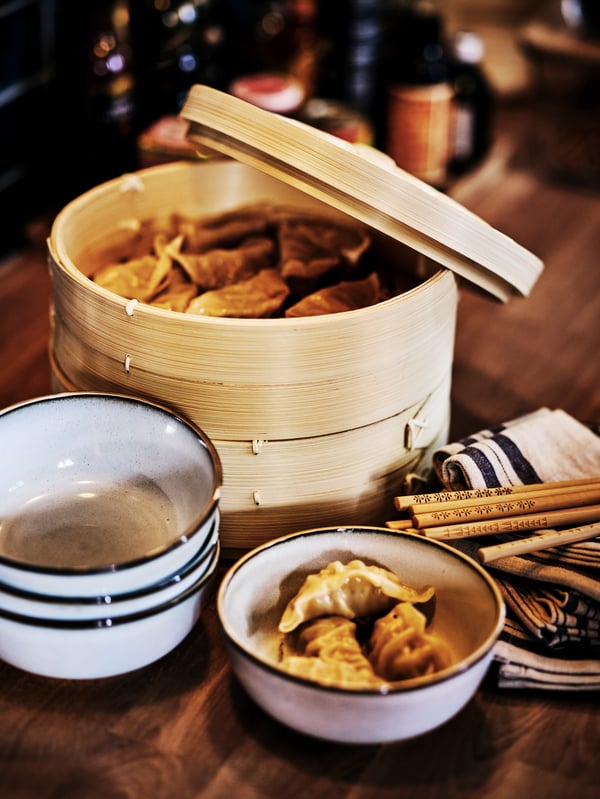 A bamboo KLOCKREN steamer filled with wantons, next to a set of grey GLADELIG bowls, chopsticks and a tea towel.