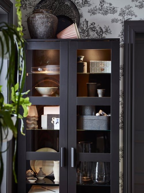 Une armoire noire avec des portes en verre BRIMNES avec des étagères en verre et un éclairage supplémentaire qui illumine les décors tels que les boîtes, les bols et les grands vases.