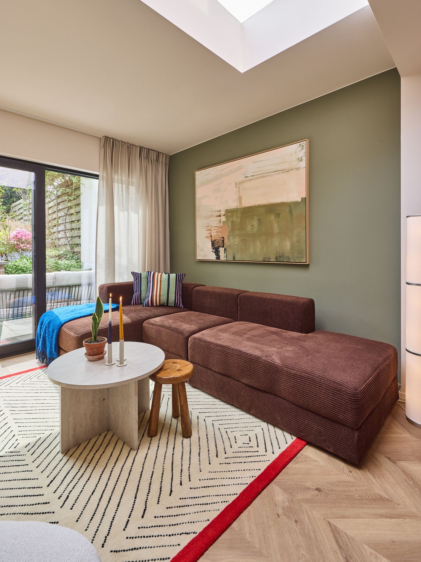Zithoek met bruine sofa, rond tafeltje en krukje op tapijt, groot schilderij aan groene muur en veel licht via raam en dakkoepel.