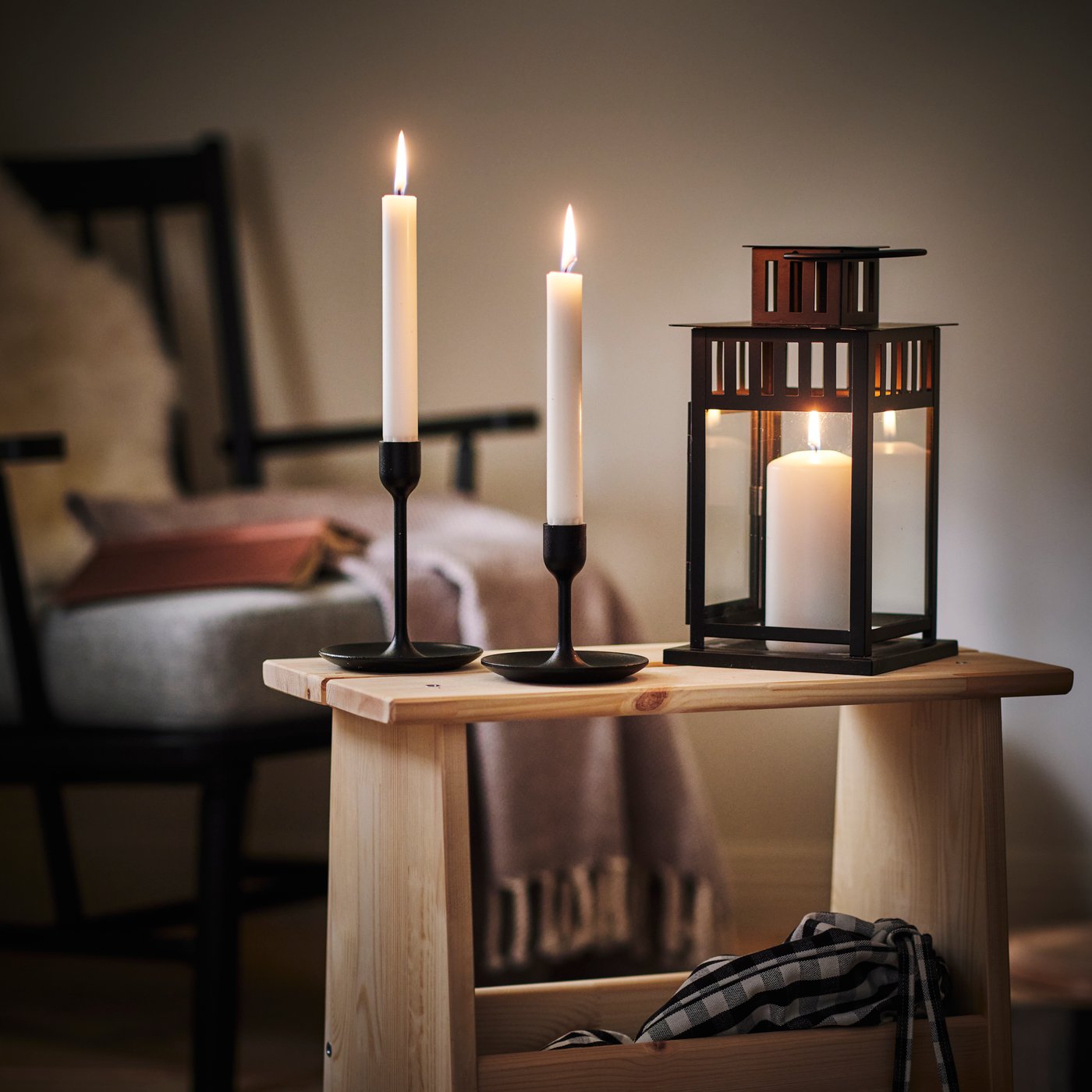 Close-up of candles in FULLTALIG candle holder and BORRBY lantern, on top of a wooden side table in a dark, cosy room.