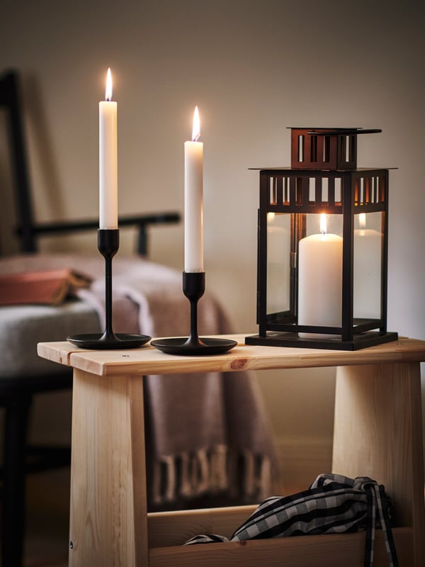 Close-up of candles in FULLTALIG candle holder and BORRBY lantern, on top of a wooden side table in a dark, cosy room.