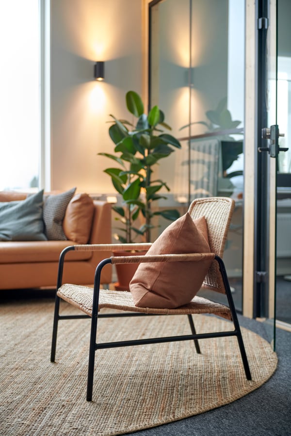 Modern och mysig loungedel med ULRIKSBERG rottingfåtölj på rund matta, brun kudde, beige soffa med kuddar i bruna och grå toner, stor grön växt, vägglampor med varmt ljus och glasdörrar som släpper in naturligt ljus.