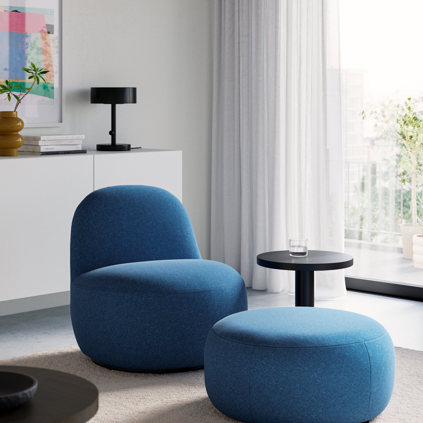 Blue upholstered accent chair with a matching round ottoman in a bright, modern living space, complemented by a small black side table and large windows.