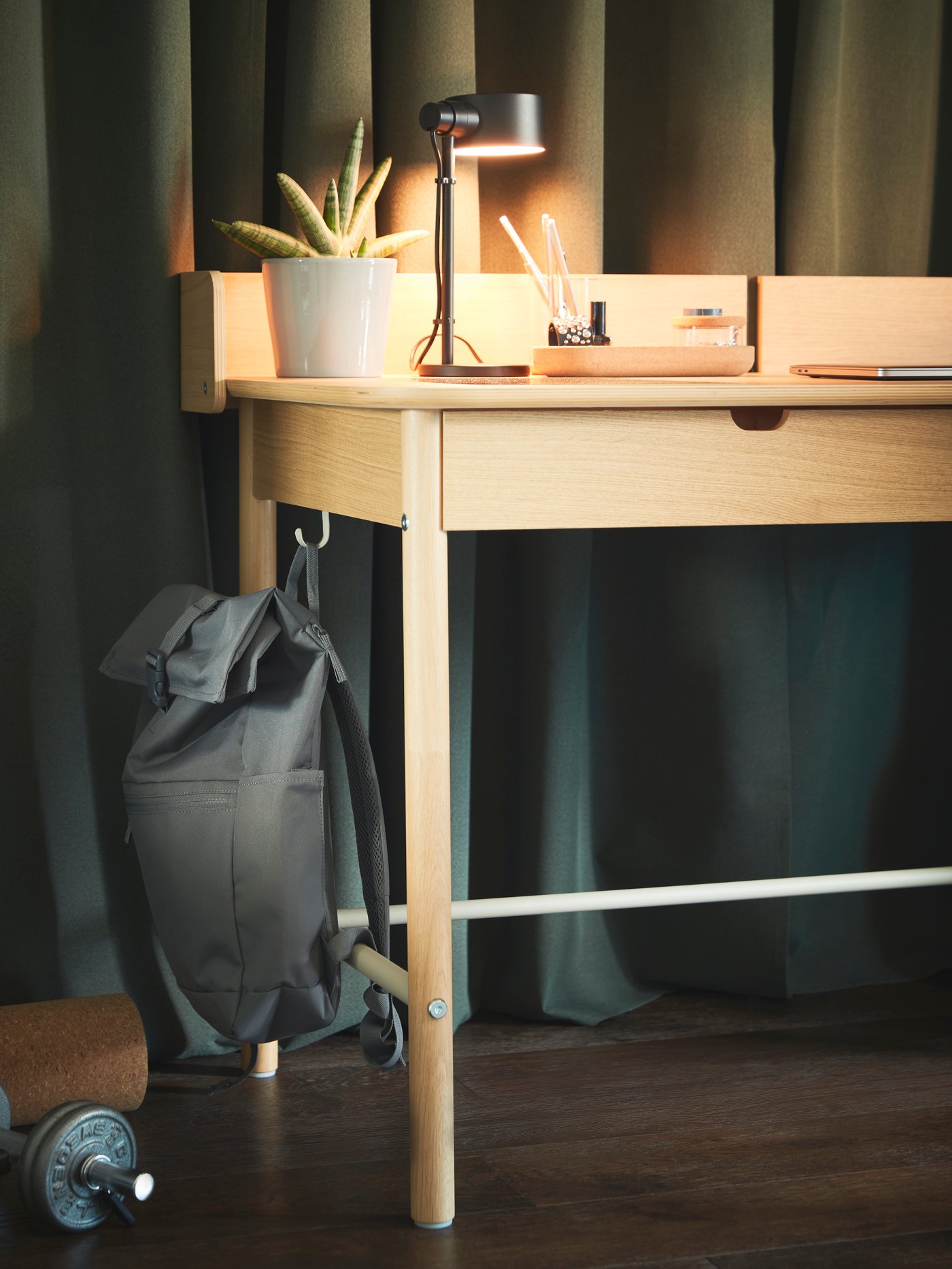 A grey STARTTID backpack hangs on the side hook of an oak-veneer RIDSPÖ desk that has a lit black LÖVMÅNAD work lamp on it.