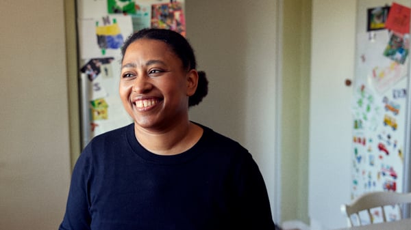 Alle in her kitchen smiling, sticker covered refrigerator and cupboards in the background.
