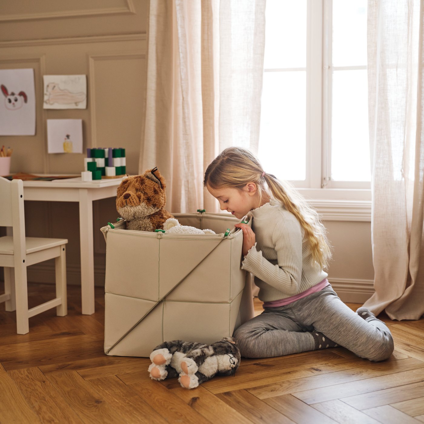 A child looks inside a GREJSIMOJS mat/storage box containing toys on the floor beside a white SUNDVIK children’s table.