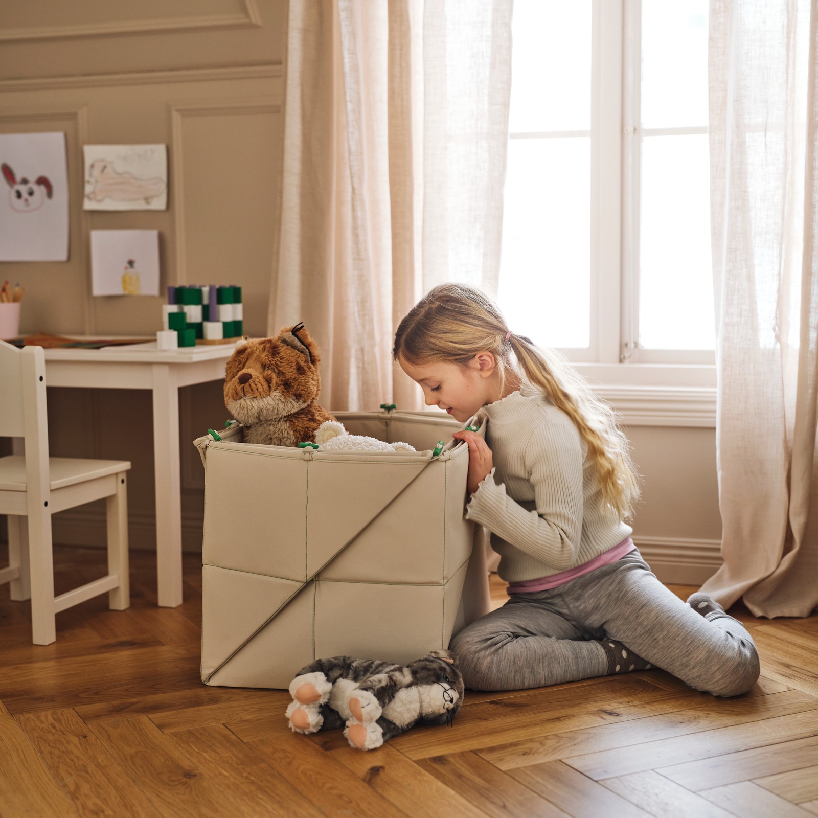 A child looks inside a GREJSIMOJS mat/storage box containing toys on the floor beside a white SUNDVIK children’s table.