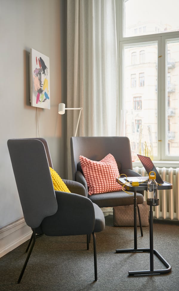 Mysig sittgrupp vid fönster med två grå BINGSTA fåtöljer, med en rosa och en gul kudde, framför ett GJÖRKÅSEN laptopbord. dekorativa kuddar i ett modernt vardagsrum.